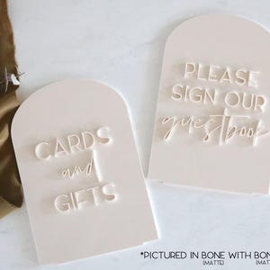 May include: Two off-white, arched signs with engraved text. One reads "CARDS and GIFTS," the other "PLEASE SIGN OUR guestbook." The signs are matte and feature bone lettering, suitable for weddings or events.