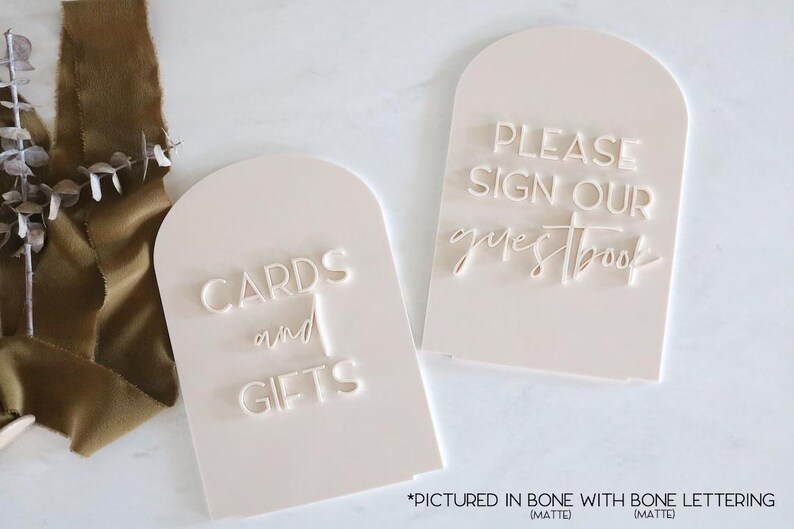 May include: Two off-white, arched signs with engraved text. One reads "CARDS and GIFTS," the other "PLEASE SIGN OUR guestbook." The signs are matte and feature bone lettering, suitable for weddings or events.