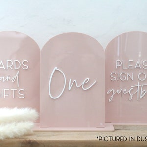 May include: Three blush pink acrylic signs with arched tops. The signs read "CARDS and GIFTS", "One", and "PLEASE SIGN OUR guestbook" in white script. The signs are for a wedding or event.