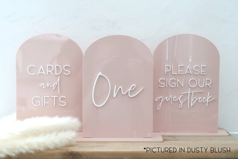 May include: Three blush pink acrylic signs with arched tops. The signs read "CARDS and GIFTS", "One", and "PLEASE SIGN OUR guestbook" in white script. The signs are for a wedding or event.