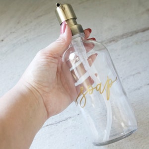 Pu&ograve; includere: Un dispenser di sapone in vetro trasparente con una pompa dorata e un'etichetta dorata con la scritta "soap" in corsivo.