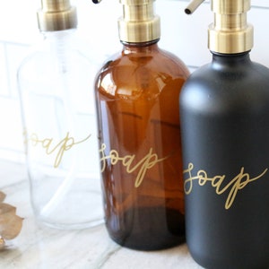 Pu&ograve; includere: Tre dispenser di sapone in vetro con pompe dorate ed etichette che recitano "sapone" in scrittura dorata. I dispenser sono trasparenti, ambrati e neri.