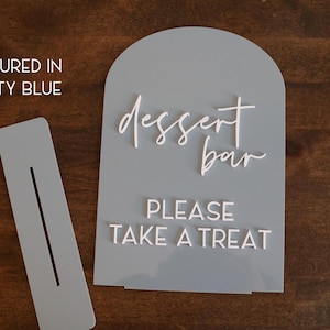 May include: A dusty blue arched acrylic sign with white 3D lettering that reads "dessert bar" in script and "PLEASE TAKE A TREAT" in block letters, accompanied by a matching rectangular base with a slot for the sign to stand upright.