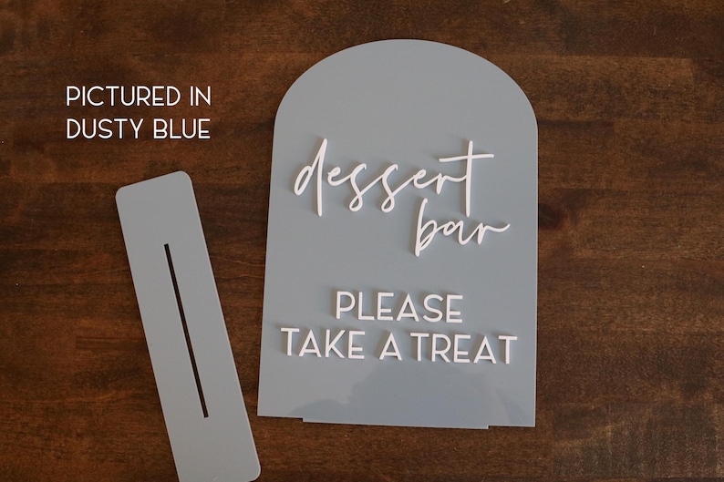 May include: A dusty blue arched acrylic sign with white 3D lettering that reads "dessert bar" in script and "PLEASE TAKE A TREAT" in block letters, accompanied by a matching rectangular base with a slot for the sign to stand upright.