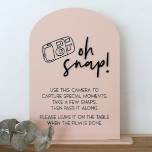 May include: A blush pink sign with an arched top and a rectangular base. The sign features a black line drawing of a camera and the words "oh snap!" in a handwritten font. Below, text instructs users to capture special moments.