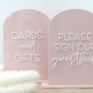 May include: Two blush pink acrylic signs with arched tops. One sign reads "CARDS and GIFTS" in white script, the other says "PLEASE SIGN OUR guestbook". Both signs are on a matching base, perfect for a wedding or event.
