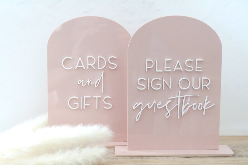 May include: Two blush pink acrylic signs with arched tops. One sign reads "CARDS and GIFTS" in white script, the other says "PLEASE SIGN OUR guestbook". Both signs are on a matching base, perfect for a wedding or event.