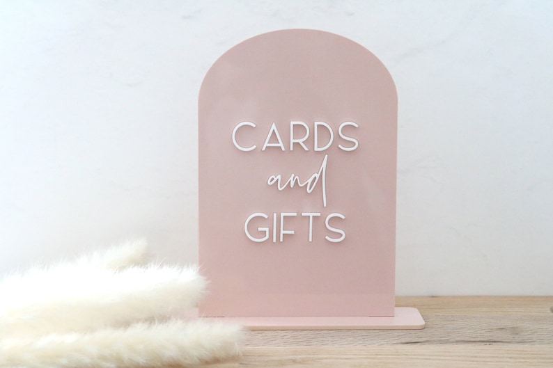 May include: A blush pink acrylic sign with an arched top, displaying the words "CARDS and GIFTS" in white, elegant script. The sign is designed to stand upright, perfect for weddings or events. The background is a soft white.