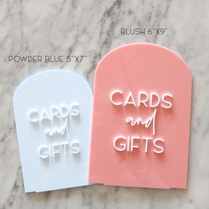 May include: Two acrylic signs with arched tops. One is powder blue, 5"x7", and the other is blush, 6"x9". Both signs have the words "CARDS and GIFTS" in white, stylized lettering. Ideal for weddings or events.