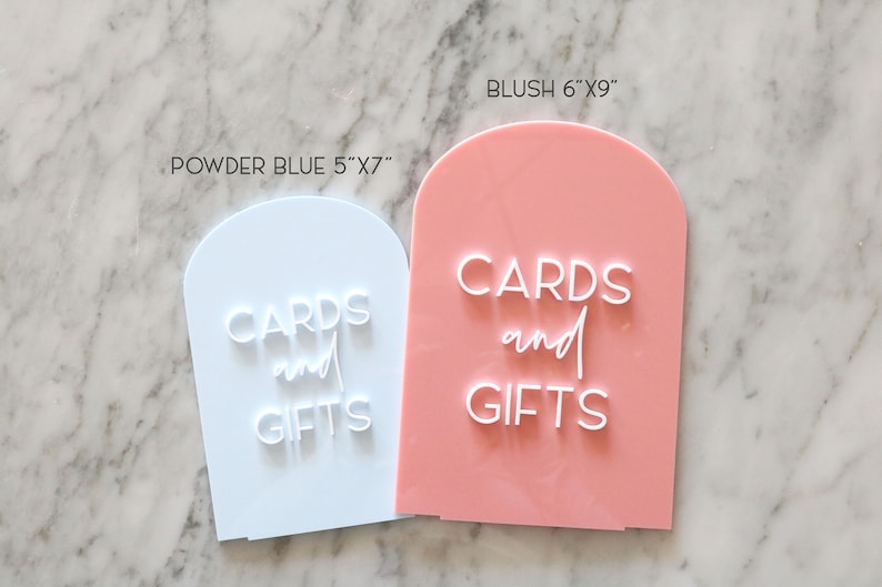 May include: Two acrylic signs with arched tops. One is powder blue, 5"x7", and the other is blush, 6"x9". Both signs have the words "CARDS and GIFTS" in white, stylized lettering. Ideal for weddings or events.
