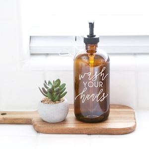 May include: A brown glass soap dispenser with a black pump top. The dispenser has the text "wash your hands" printed on it in white. The dispenser is sitting on a wooden cutting board with a small potted succulent plant.