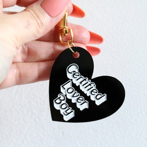 May include: Black heart-shaped keychain with a gold clasp. The keychain has white text that reads "Certified Lover Boy".