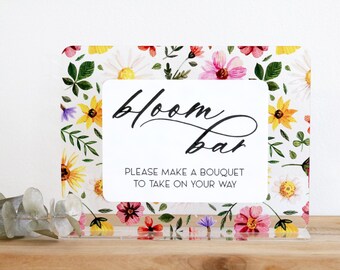 May include: A clear acrylic sign with a floral design and the words "bloom bar." The sign has a white background with colorful flowers and greenery. The text "PLEASE MAKE A BOUQUET TO TAKE ON YOUR WAY" is also included.