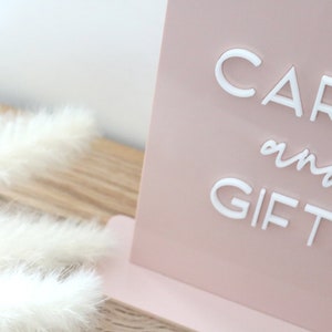 May include: A blush pink sign with the words "CARDS and GIFTS" in white, elegant lettering. The sign is propped up on a matching pink base. Fluffy white feathers are in the foreground.