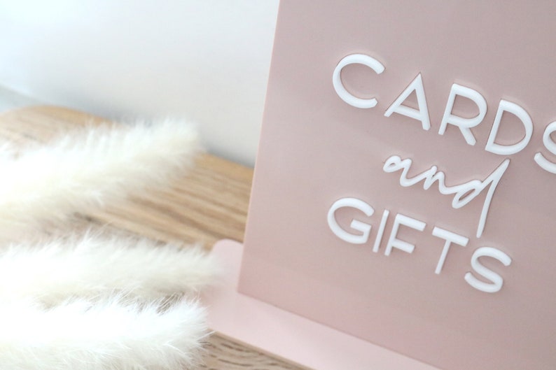 May include: A blush pink sign with the words "CARDS and GIFTS" in white, elegant lettering. The sign is propped up on a matching pink base. Fluffy white feathers are in the foreground.