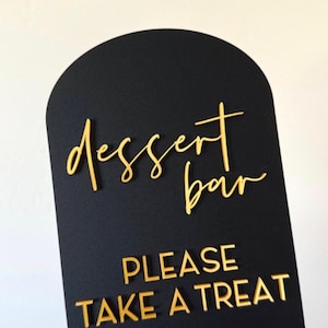 May include: Black acrylic dessert bar sign with gold metallic lettering. The sign reads "dessert bar" in cursive and "PLEASE TAKE A TREAT" in block letters. The top of the sign is arched.