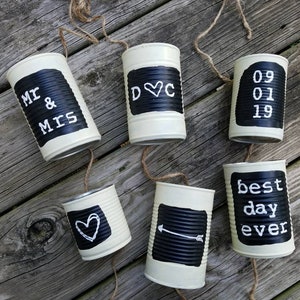 May include: A string of six white painted tin cans with black chalkboard labels. The labels read "Mr & Mrs", "D&C", "09 01 19", "best day ever", a heart, and an arrow. The cans are strung together with twine.