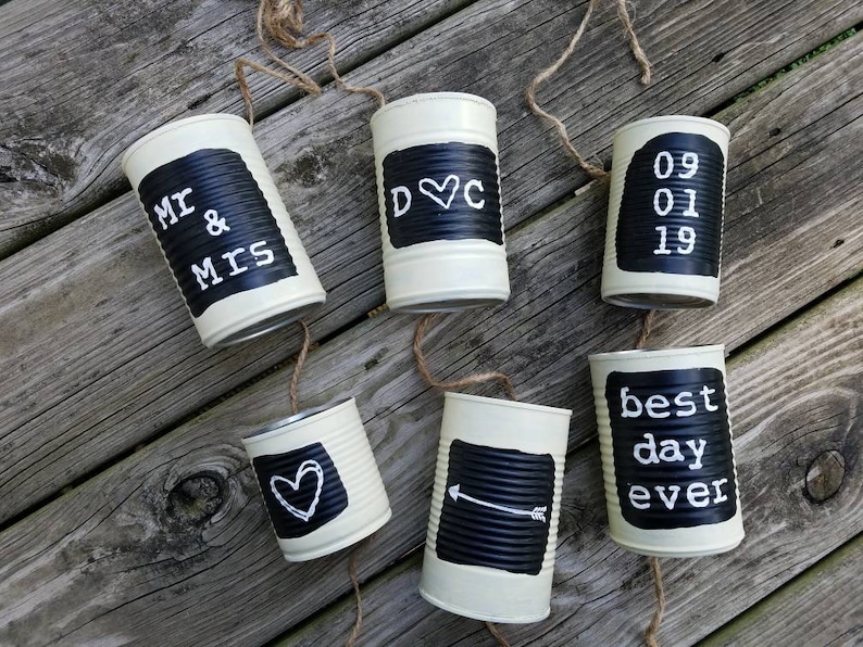 May include: A string of six white painted tin cans with black chalkboard labels. The labels read "Mr & Mrs", "D&C", "09 01 19", "best day ever", a heart, and an arrow. The cans are strung together with twine.
