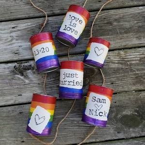 May include: Six rainbow painted cans with string attached. The cans have various messages written on them, including "Love is Love", "Just Married", "Sam & Nick", and "13-20".