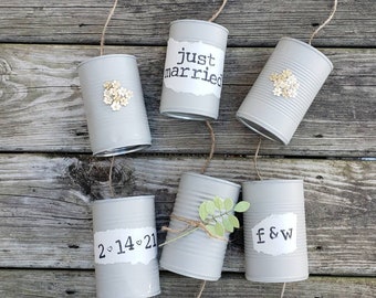 Op de afbeelding: Een reeks van zes grijs geverfde blikken met touw eraan bevestigd. De blikken zijn versierd met papier, bloemen, groen en de tekst "just married", "2-14-21" en "F&W".