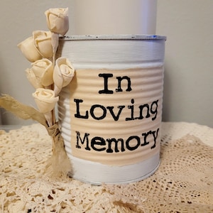 May include: A white painted tin can with the words "In Loving Memory" printed on a beige label. A bundle of cream-colored artificial roses is tied to the side with a beige ribbon. The can sits on a cream-colored lace doily.