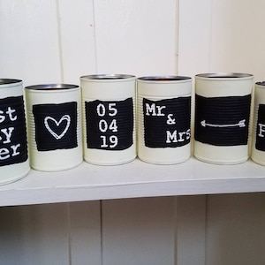 May include: Six white painted cans with black chalkboard labels. The labels have the following text: "best day ever", a heart, "05 04 19", "Mr & Mrs", an arrow, and "F &hearts; W".