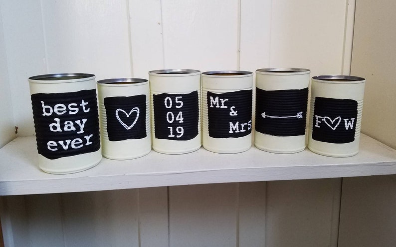 May include: Six white painted cans with black chalkboard labels. The labels have the following text: "best day ever", a heart, "05 04 19", "Mr & Mrs", an arrow, and "F &hearts; W".