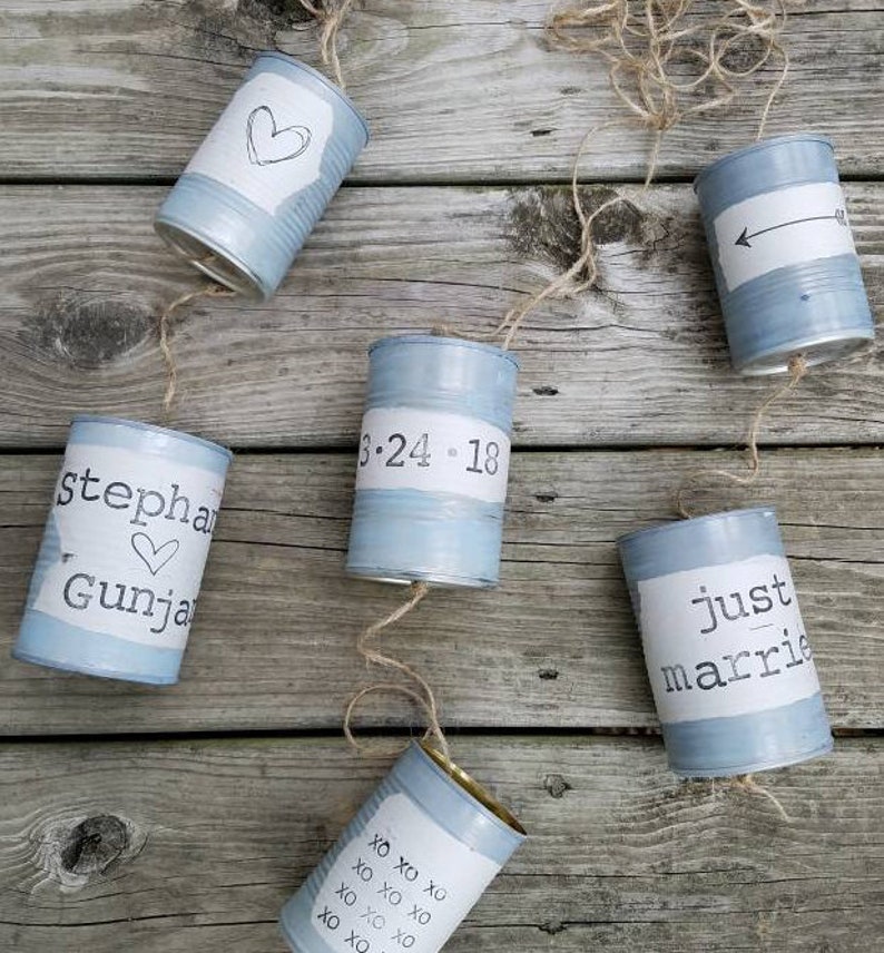 Op de afbeelding: Een reeks van zes blauwe beschilderde blikken met witte letters. De blikken zijn versierd met harten, pijlen, de datum 3-24-18, de woorden "stepha Gunja", "just marrie" en "XO XO XO OX OX OX OX OX XO XO".
