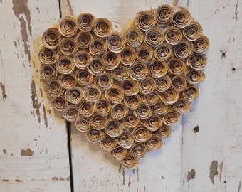 Op de afbeelding: Een hartvormige wanddecoratie gemaakt van papier, met een rustieke, vintage uitstraling. Het hart is gemaakt van veel kleine, gerolde papieren bloemen, allemaal in tinten bruin en beige.