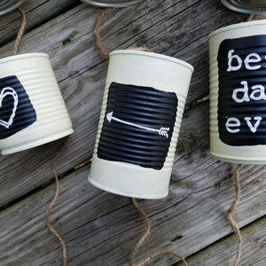 May include: Three white painted tin cans with black chalkboard paint and white lettering. The cans are strung together with twine. The text on the cans reads "best day ever", a heart, and an arrow.