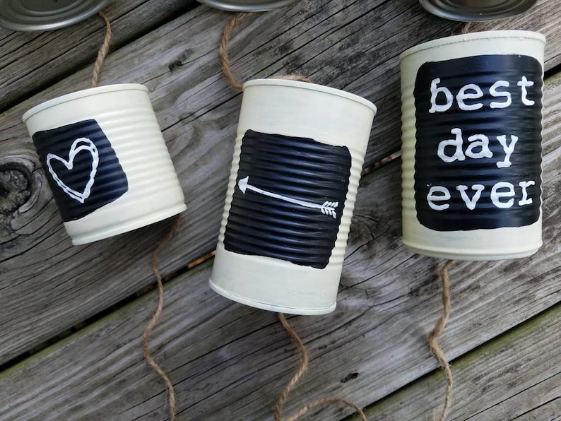 May include: Three white painted tin cans with black chalkboard paint and white lettering. The cans are strung together with twine. The text on the cans reads "best day ever", a heart, and an arrow.