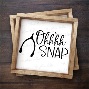 May include: A wooden sign with a white background and black text that reads "Ohhhh SNAP" with a black horseshoe-shaped good luck charm to the left of the text.