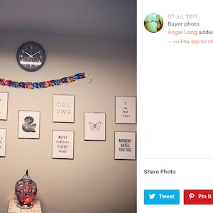 May include: A white wall with a variety of framed prints featuring inspirational quotes and designs. The prints include phrases like "Be Nice or Leave", "You Got This", "Girl Power", "Good Vibes Only", and "Monday Hates You Too". A clock is hanging on the wall, and a decorative birdcage with red and orange accents is on a table in the foreground.