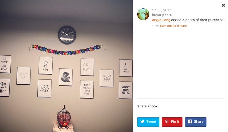 May include: A white wall with a variety of framed prints featuring inspirational quotes and designs. The prints include phrases like "Be Nice or Leave", "You Got This", "Girl Power", "Good Vibes Only", and "Monday Hates You Too". A clock is hanging on the wall, and a decorative birdcage with red and orange accents is on a table in the foreground.