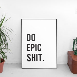 Puede incluir: Un p&oacute;ster enmarcado en negro con fondo blanco y el texto "DO EPIC SHIT." en letras negras. El p&oacute;ster est&aacute; apoyado contra una pared blanca. Una linterna verde y una maleta marr&oacute;n est&aacute;n sobre una superficie blanca en primer plano.