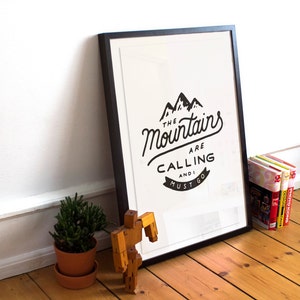 May include: A black framed print with a white background and black text that reads "The Mountains Are Calling And I Must Go". The print is hanging on a white wall with a wooden floor.