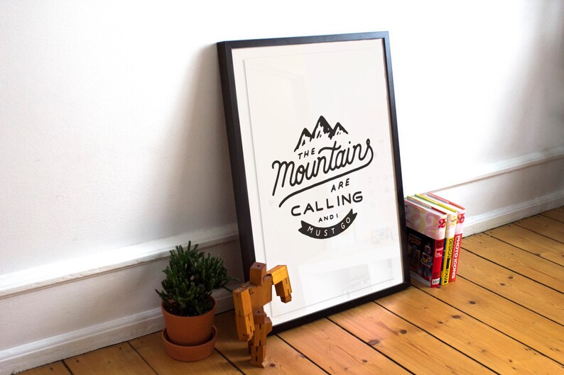 May include: A black framed print with a white background and black text that reads "The Mountains Are Calling And I Must Go". The print is hanging on a white wall with a wooden floor.
