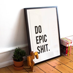 Puede incluir: Un p&oacute;ster enmarcado en negro con el texto "DO EPIC SHIT." sobre un fondo blanco. El p&oacute;ster est&aacute; apoyado en una pared blanca y hay una peque&ntilde;a planta en maceta y un juguete de madera en primer plano.
