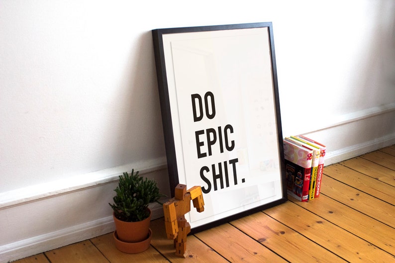 Puede incluir: Un p&oacute;ster enmarcado en negro con el texto "DO EPIC SHIT." sobre un fondo blanco. El p&oacute;ster est&aacute; apoyado en una pared blanca y hay una peque&ntilde;a planta en maceta y un juguete de madera en primer plano.