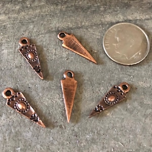 May include: Close-up of five copper-colored metal charms. Four charms are triangular with decorative patterns and a loop at the top. One charm is a simple triangle. A dime is included for size reference. The charms are on a gray surface.