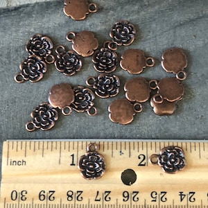 May include: A collection of 15 copper-colored metal flower charms with a loop for attaching to jewelry or other crafts. The charms are approximately 1 inch in diameter and feature a detailed floral design.