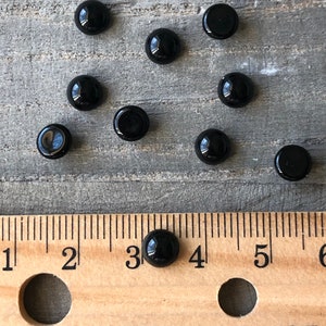 May include: Ten black glass cabochons, approximately 3/8 inch in diameter, arranged in a pattern on a wooden surface with a ruler for scale.
