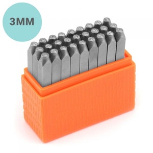 May include: A set of 3mm metal letter stamps in an orange plastic storage box. The stamp heads are visible, and the text "3MM" is in a light blue circle in the upper left corner. The stamps are arranged neatly.