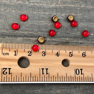 May include: A close-up of red and gold round glass cabochons, measuring approximately 3/8 inch in diameter, scattered on a wooden surface with a ruler for scale.