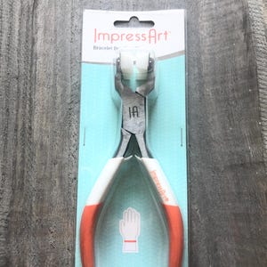 May include: A pair of red and white pliers with a silver jaw and a white plastic piece in the jaw. The pliers are labeled "ImpressArt" and "Jewelry Tool".