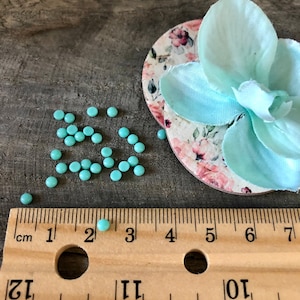 May include: A close-up of a ruler measuring 7 inches with a single turquoise bead on the 3-inch mark. Scattered around the ruler are approximately 30 turquoise beads.