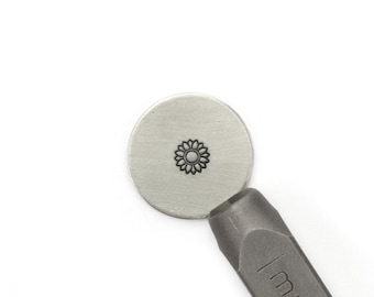 May include: A close-up of a metal stamp with a sunflower design. The stamp is being used to create an impression on a metal disc.