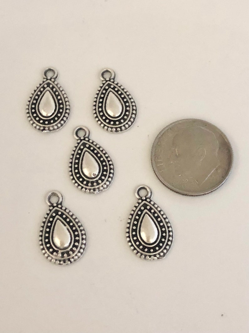 May include: Five silver-colored teardrop charms with a dotted border and a loop at the top. A dime is next to the charms for size comparison. The charms have a simple, elegant design.