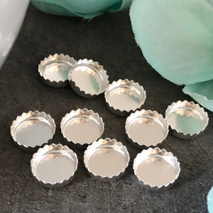 May include: Ten silver-colored metal bottle caps with a scalloped edge. The caps are arranged in a semi-circle on a gray surface.