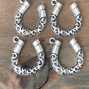 May include: Four silver-toned metal horseshoe-shaped charms with an ornate floral design. The charms have a loop at the top for attaching to jewelry or other crafts.